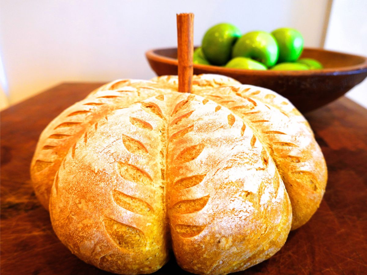 a pumpkin shaped sourdough bread decorated with scoring sitting on a butcher block table with a bowl of apples